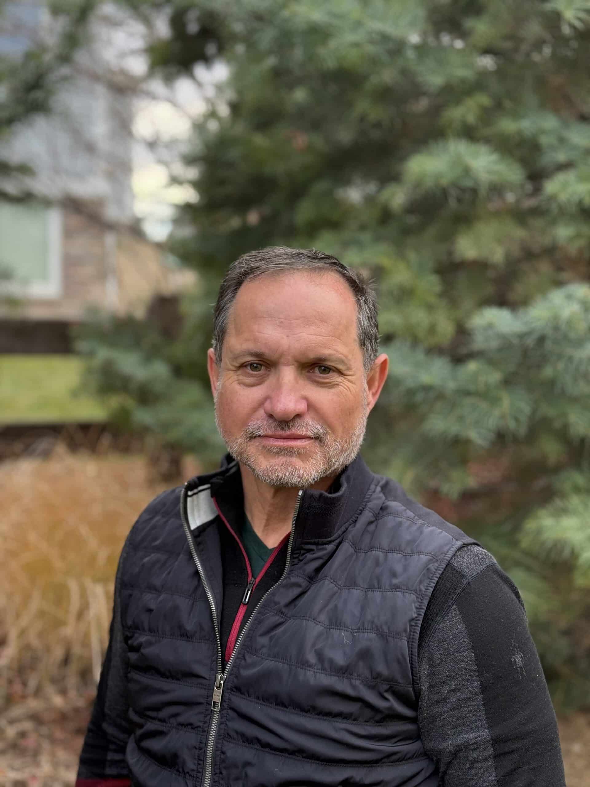 Man with salt and pepper hair wearing a black vest in front of a green tree.