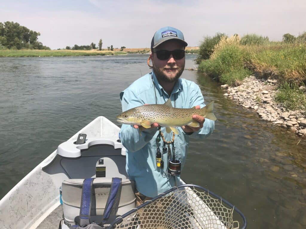 Man holding a fish in a boat on a sunny river, wearing sunglasses and blue shirt, showcasing catch while fishing.