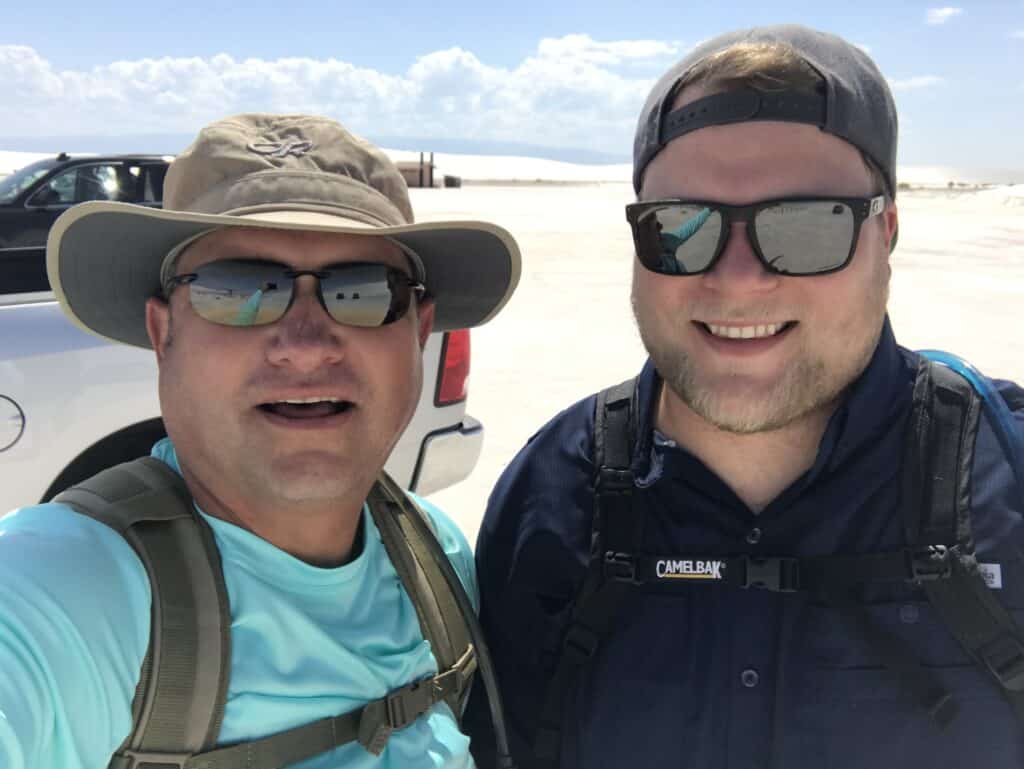 Two men in sunglasses smiling at a sunny desert landscape with a parked vehicle in the background.